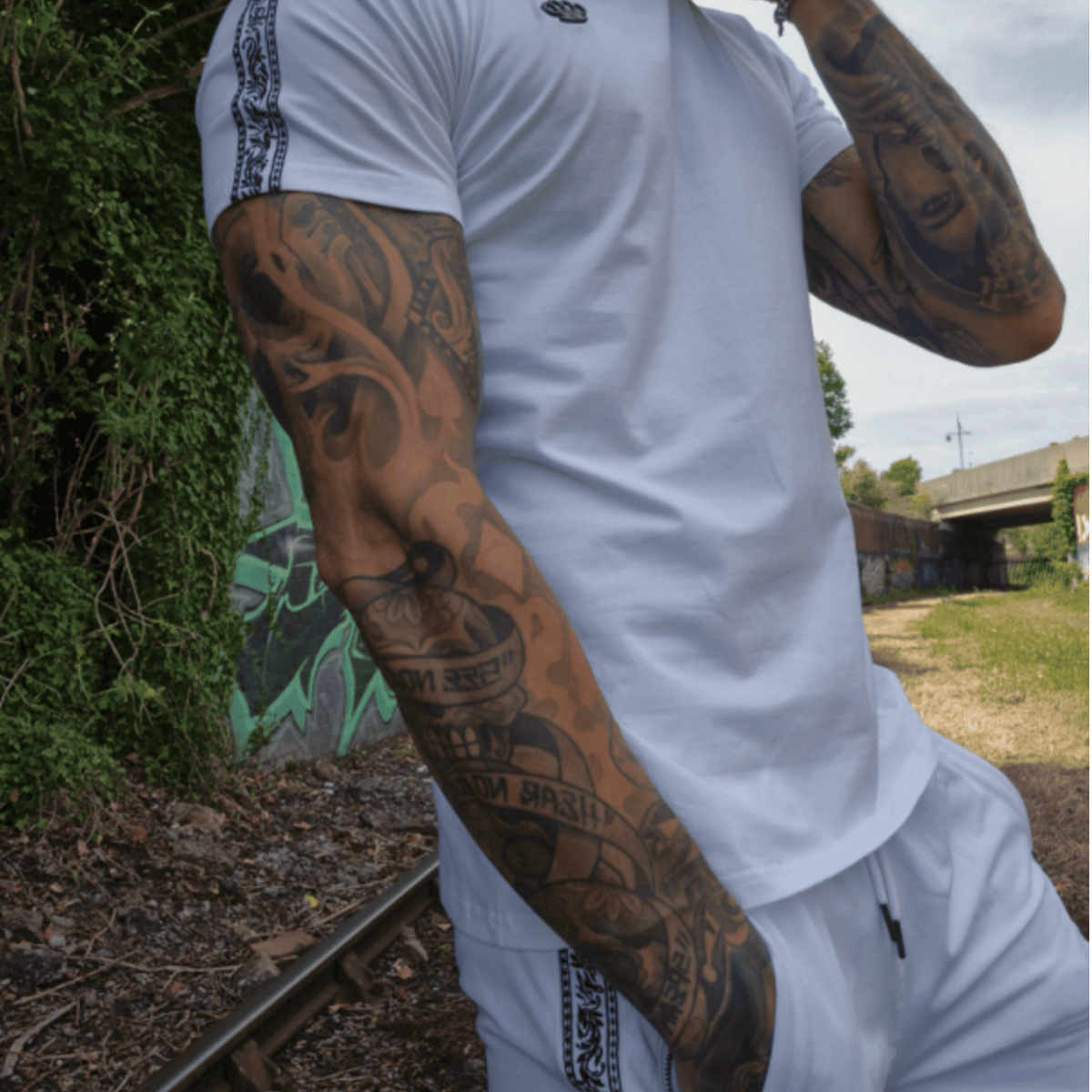 Model in a white MVL T-shirt and white Meander shorts by railway tracks with graffiti